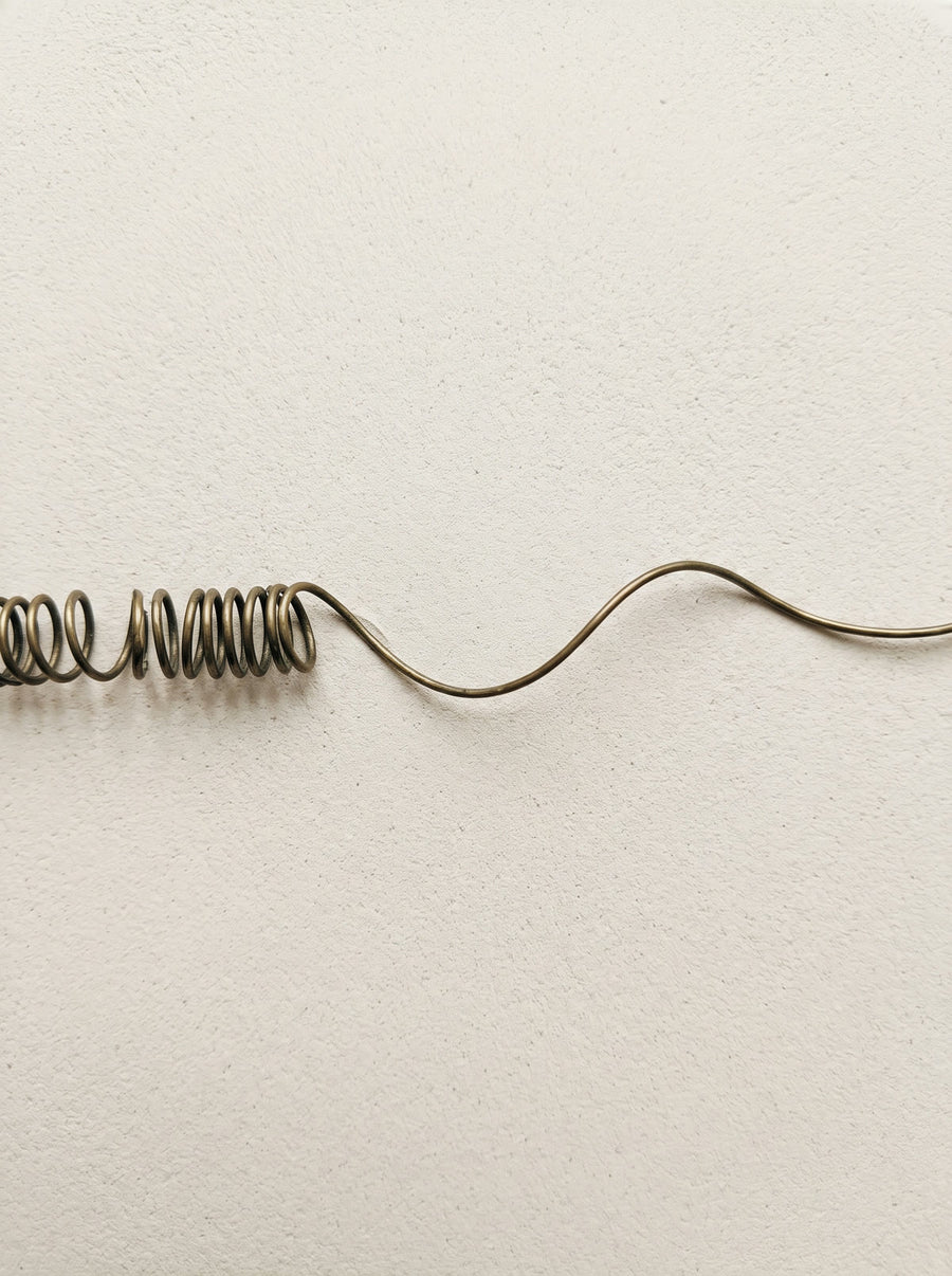 A bronze wire uncoiling from a tight coil into an open wave across pale plaster