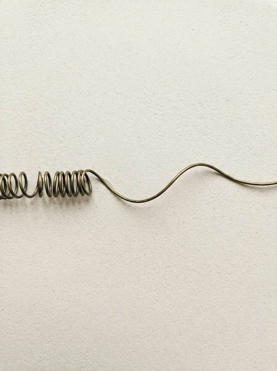 A coiled bronze wire unwinding into a slow wave across pale plaster